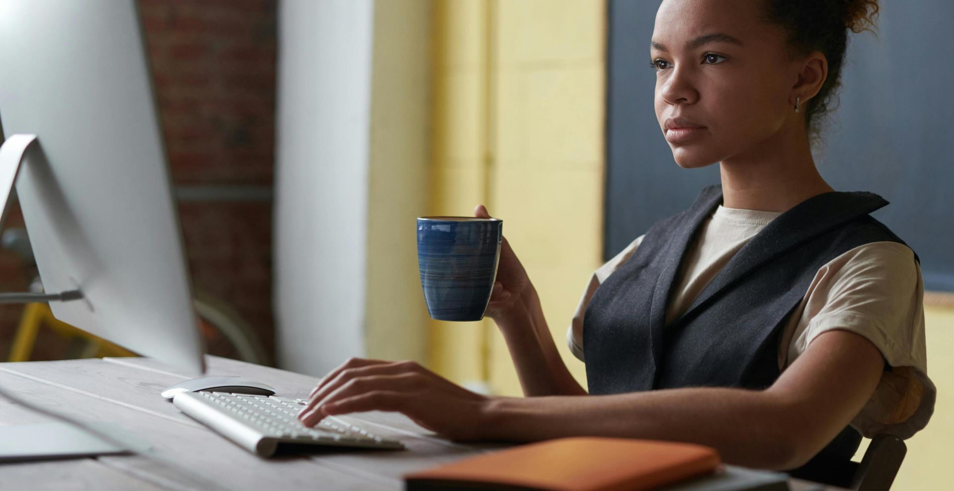 A professional young woman concentrating on work at a desk with a computer and coffee cup in hand.